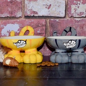 May include: Two ceramic cat-shaped pet food bowls with raised legs. The yellow bowl says "Diesel" and the gray bowl says "Harley".