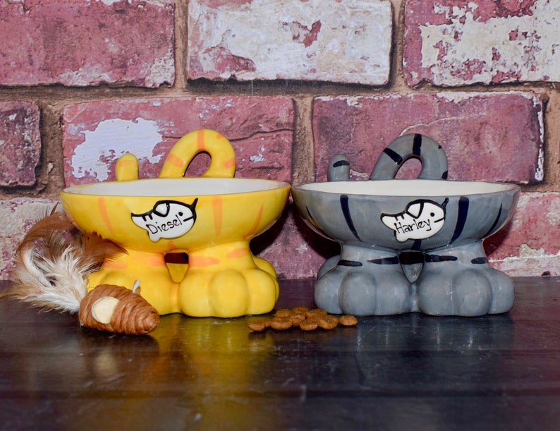 May include: Two ceramic cat-shaped pet food bowls with raised legs. The yellow bowl says "Diesel" and the gray bowl says "Harley".