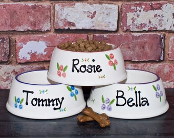 May include: Three white ceramic dog bowls with blue, pink, and purple floral designs. The bowls are stacked with the top bowl filled with brown dog food. The bowls are personalized with the names "Rosie", "Tommy", and "Bella".