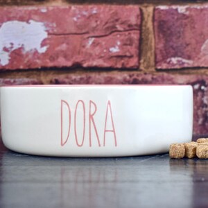 K&ouml;nnte beinhalten: Wei&szlig;e Keramik-Futternapf f&uuml;r Haustiere mit rosa Rand und dem Namen "DORA" in rosa Schrift. Die Sch&uuml;ssel steht auf einer dunklen Holzoberfl&auml;che mit ein paar braunen Hundekeksen davor.