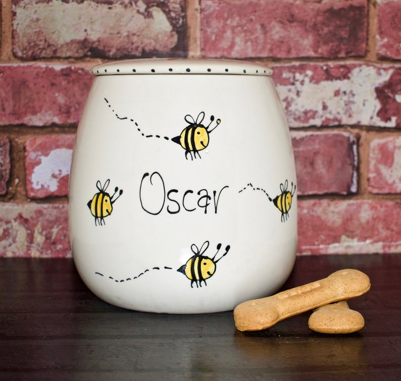May include: A white ceramic treat jar with a lid, featuring hand-painted yellow and black bees flying along dotted flight paths. The name Oscar is written in black script in the center. Two light brown bone-shaped dog treats sit in front.