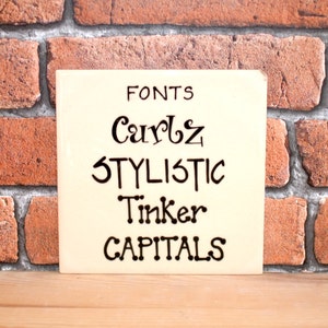 May include: A white ceramic tile with black lettering that reads "FONTS", "Curls", "STYLISTIC", "Tinker", and "CAPITALS".