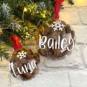 May include: Two clear baubles filled with dog treats. One has the name "Bailey" in white script, the other "Luna". Both have a white snowflake and a red ribbon with white polka dots for hanging. Festive decorations for pets.