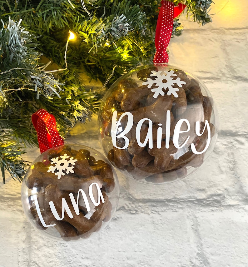 May include: Two clear baubles filled with dog treats. One has the name "Bailey" in white script, the other "Luna". Both have a white snowflake and a red ribbon with white polka dots for hanging. Festive decorations for pets.