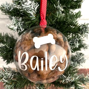 May include: A clear, spherical Christmas ornament filled with dog biscuits. The ornament has the name "Bailey" in white script and a white bone shape. It hangs from a red ribbon with white spots, against a green Christmas tree.