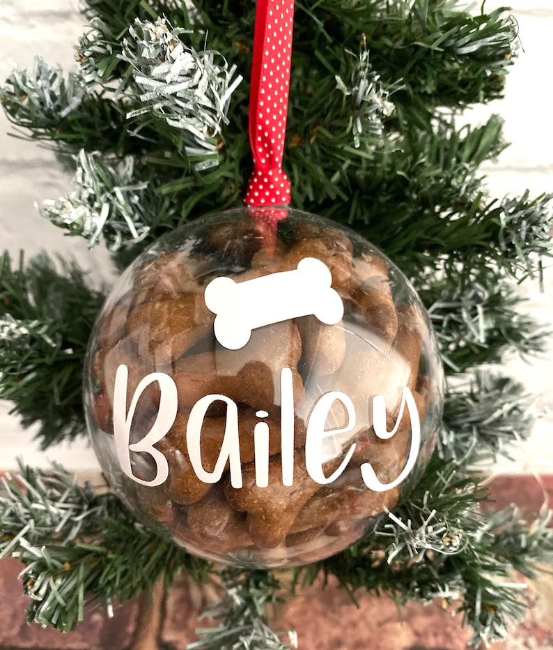 May include: A clear, spherical Christmas ornament filled with dog biscuits. The ornament has the name "Bailey" in white script and a white bone shape. It hangs from a red ribbon with white spots, against a green Christmas tree.