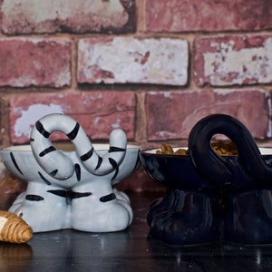 May include: Two ceramic pet food bowls shaped like cats. The bowl on the left is gray and white with black stripes and the bowl on the right is dark blue. Both bowls have food in them.