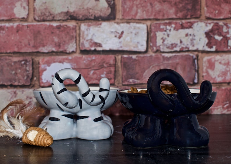 May include: Two ceramic pet food bowls shaped like cats. The bowl on the left is gray and white with black stripes and the bowl on the right is dark blue. Both bowls have food in them.