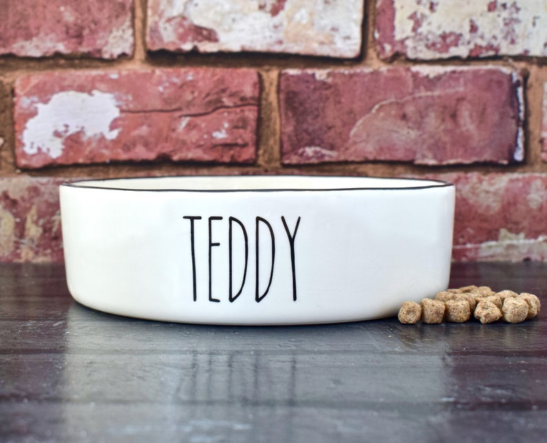 K&ouml;nnte beinhalten: Wei&szlig;e Keramik-Hunde- oder Katzennapf mit schwarzem Rand und dem Namen "TEDDY" in schwarzer Schrift auf der Vorderseite. Im Vordergrund befindet sich ein kleiner Haufen Hundefutter.