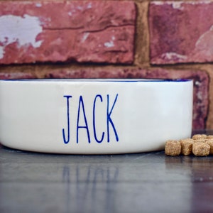 K&ouml;nnte beinhalten: Wei&szlig;e Keramik-Hundenapf mit blauem Rand und dem Namen "JACK" in blauer Schrift. Im Vordergrund liegen ein paar Hundefutter.