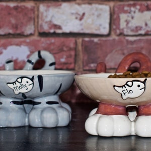May include: Two ceramic cat-shaped pet food bowls. The bowl on the left is gray with black stripes and the name "Molly" written on it. The bowl on the right is beige with brown stripes and the name "Flo" written on it.