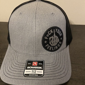 May include: A gray and black trucker hat with a black mesh back. The hat has a black patch on the front that says "F*CK YOUR FEELINGS" with a skeleton hand giving the middle finger. The hat is adjustable and has a Richardson 112 label on the side.
