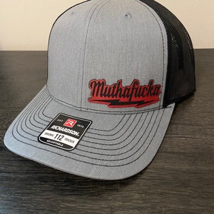 May include: A gray and black trucker hat with a red embroidered patch that says "Muthafucka". The hat is adjustable and has a Richardson 112 label.