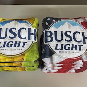 May include: Two cornhole bags. One bag features a design with the text "BUSCH LIGHT" in blue and white, with a mountain and sky graphic, and the words "BREWED IN U.S.A." The other bag has a red, white, and blue American flag design.