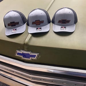 May include: Three gray and black baseball caps with a brown leather Chevrolet logo on the front. The caps are sitting on the hood of a green truck.