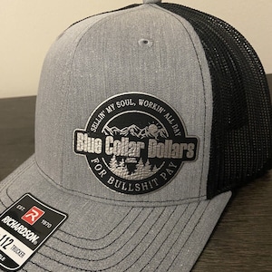 May include: A gray and black trucker hat with a silver patch that reads "Sellin' my soul, workin' all day. Blue Collar Dollars for Bullshit Pay". The hat has a mesh back and a Richardson logo on the side.