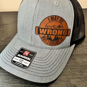 May include: Gray trucker hat with a black mesh back. The hat features a brown leather patch with the text "I MAY BE WRONG BUT I DOUBT IT" and a mountain and tree design. The hat has a Richardson tag.