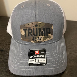 May include: A gray and white trucker hat with a camouflage patch that says "TRUMP 45 47". The hat is adjustable and has the Richardson logo on the side.