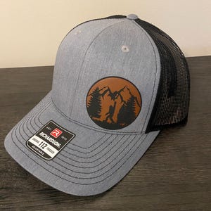 May include: A gray and black trucker hat with a brown leather patch featuring a silhouette of a person hiking in a mountain landscape. The patch has the text "112" on it. The hat is adjustable and has the Richardson brand logo on the side.