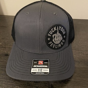 May include: A gray and black trucker hat with a black patch that says "F*ck Your Feelings" in white lettering. The hat is adjustable and has a Richardson logo on the side.