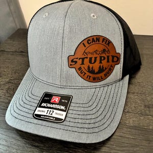 May include: Gray trucker hat with a black mesh back. The hat features a brown leather patch with the text "I CAN FIX STUPID BUT IT WILL HURT" and a mountain and tree graphic. The hat is adjustable.