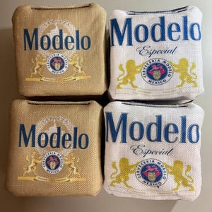 May include: Four square bean bags with the Modelo logo. Two bags are tan with blue text and a gold emblem. The other two are white with blue text and the words "Especial" and a gold emblem.