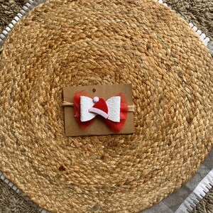 May include: A red and white Christmas-themed hair bow with a small red Santa hat in the center. The bow is made of tulle and glitter and is attached to a headband.