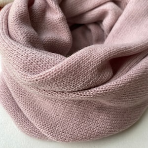 以下が含まれることがあります： 柔らかくピンク色のニットのインフィニティスカーフ。スカーフは細かいニット生地で作られており、円形に折り畳まれています。