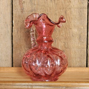 Pode incluir: Um jarro de vidro rosa com uma borda com babados e uma alça transparente. O jarro tem um corpo bulboso com um design texturizado e canelado. O jarro é exibido em uma prateleira de madeira contra um fundo de madeira.