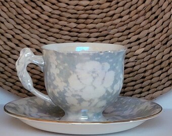 May include: A vintage teacup and saucer set. The cup and saucer are white with a light blue floral pattern. The cup has a unique handle design. The rim of the cup and saucer are trimmed with gold.