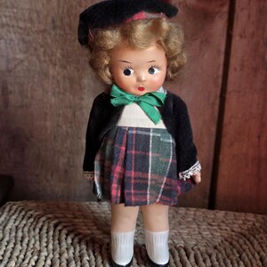 Puede incluir: Muñeca vintage con boina negra con detalle rojo, chaqueta negra y falda a cuadros en tonos rojos, verdes y azules. La muñeca tiene pelo rubio rizado, pajarita verde y calcetines blancos.
