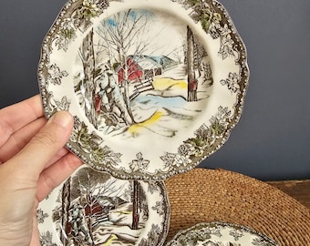 May include: Three vintage ceramic plates with a winter scene featuring a red barn, trees, and a snowy landscape. The plates have a brown and green floral border.