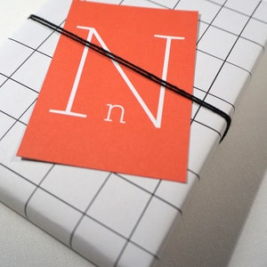 Puede incluir: Una caja de regalo blanca con un patr&oacute;n de cuadr&iacute;cula negra. La caja est&aacute; atada con una cuerda negra y tiene una tarjeta roja con una letra blanca "N" y la palabra "n" en ella.