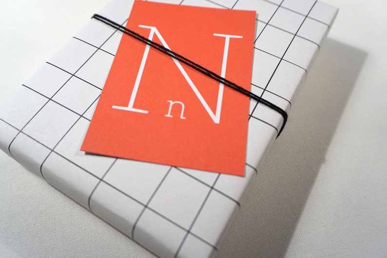 Puede incluir: Una caja de regalo blanca con un patr&oacute;n de cuadr&iacute;cula negra. La caja est&aacute; atada con una cuerda negra y tiene una tarjeta roja con una letra blanca "N" y la palabra "n" en ella.