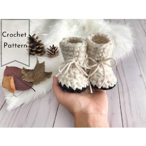 Op de afbeelding: Een paar gehaakte babyschoentjes in wit met zwarte afwerking. De schoentjes hebben een simpel ontwerp en zijn gebonden met een wit lint.