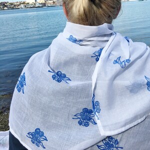 May include: A white cotton scarf with a blue floral print. The scarf is draped over a person's shoulders.