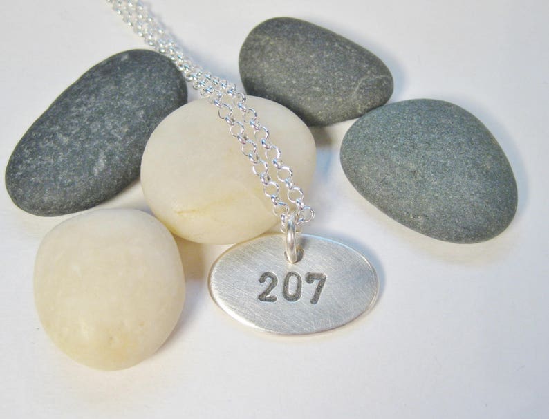 Może przedstawiać: Srebrny naszyjnik z okrągłym wisiorkiem grawerowanym numerem 207. Naszyjnik jest prezentowany na białym tle z szarymi kamieniami.