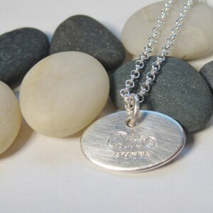 Może przedstawiać: Srebrny naszyjnik z okrągłym wisiorem, na kt&oacute;rym widnieje napis "Ciao" i "Sterling". Naszyjnik leży na białym tle z szarymi kamieniami.