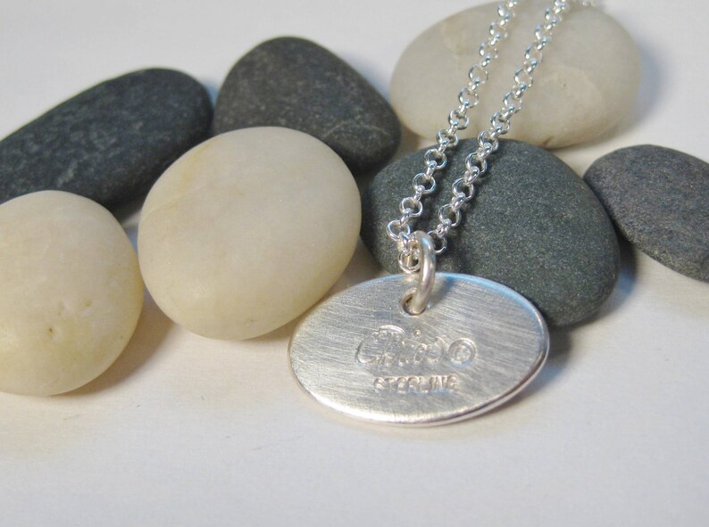 Może przedstawiać: Srebrny naszyjnik z okrągłym wisiorem, na kt&oacute;rym widnieje napis "Ciao" i "Sterling". Naszyjnik leży na białym tle z szarymi kamieniami.
