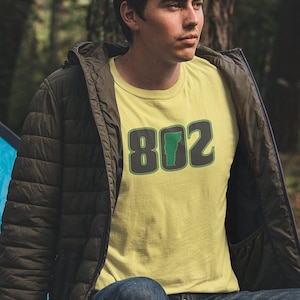 May include: A person wearing a yellow t-shirt with a green and black graphic of the state of Vermont and the area code 802. The person is sitting on a log in a forest setting.