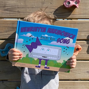 May include: A child holds a book titled "Henrietta Harmonica finds her song" with a cartoon harmonica on the cover. The harmonica has a purple bow, blue eyes, and a white face. The book has a blue background with white clouds and musical notes.