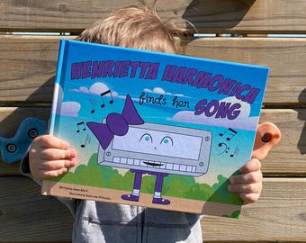 May include: A child holds a book titled "Henrietta Harmonica finds her song" with a cartoon harmonica on the cover. The harmonica has a purple bow, blue eyes, and a white face. The book has a blue background with white clouds and musical notes.