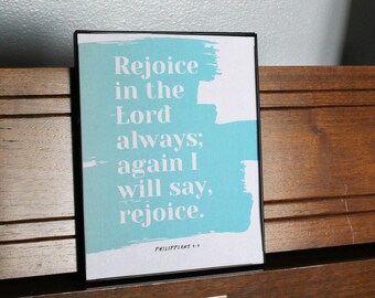 May include: A framed print with a blue background and white text that reads "Rejoice in the Lord always; again I will say, rejoice." The text is centered on the print and is surrounded by a blue brushstroke. The bottom of the print reads "Philippians 4:4".