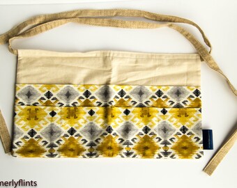 May include: A beige canvas utility apron with a geometric pattern in shades of yellow, gray, and black. The apron has a long, adjustable strap made of burlap.