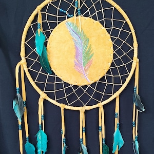 May include: A handcrafted dreamcatcher with a circular design. The center features a tan circle with a colorful feather design. Turquoise and black feathers hang from the bottom, and the frame is wrapped in tan leather.