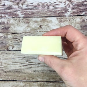 May include: A rectangular bar of white soap, held in a hand against a woodgrain background.