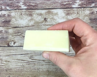 May include: A rectangular bar of white soap, held in a hand against a woodgrain background.