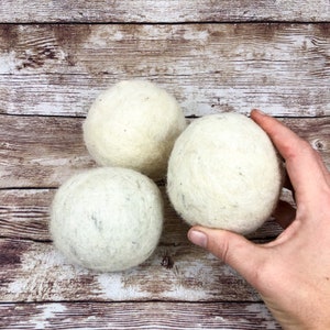 May include: Three white wool dryer balls on a wooden surface. The balls are round and have a slightly textured surface.