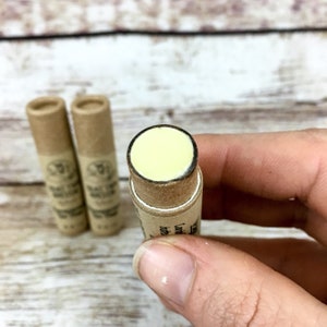 May include: Three cardboard tubes containing lip balm. The tube in the foreground is held in a hand and shows a yellow lip balm inside.