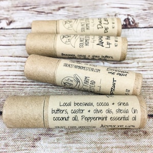 May include: Four brown cardboard tubes with labels containing information about the lip balm products. The labels include the name of the product, ingredients, and instructions for use. The products are made by Solace Farm Homestead.